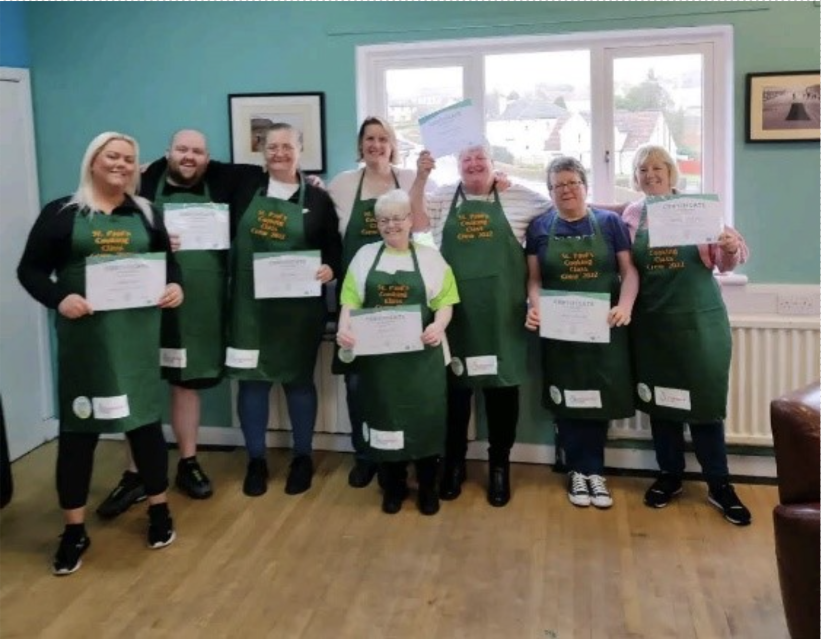 A group photo of the cooking crew wearing their aprons and holding certificates. All the people in the photo have a light skin tone.