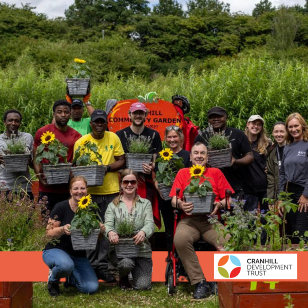 A group of people with different skin tones, some kneeling, some standing and one person is seated in a wheelchair. Many are holding potted sunflowers. Logo: Cranhill Development Trust