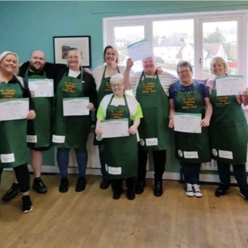 A group photo of the cooking crew wearing their aprons and holding certificates. All the people in the photo have a light skin tone.