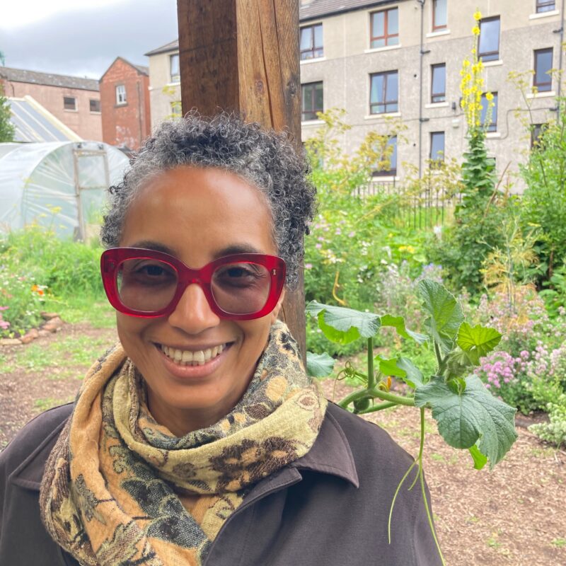 Satya, a woman with medium dark skin tone and black tighly curled hair with grey streaks stands in a luscious food garden, smiling at the camera. She wear bright red rimmed glasses.